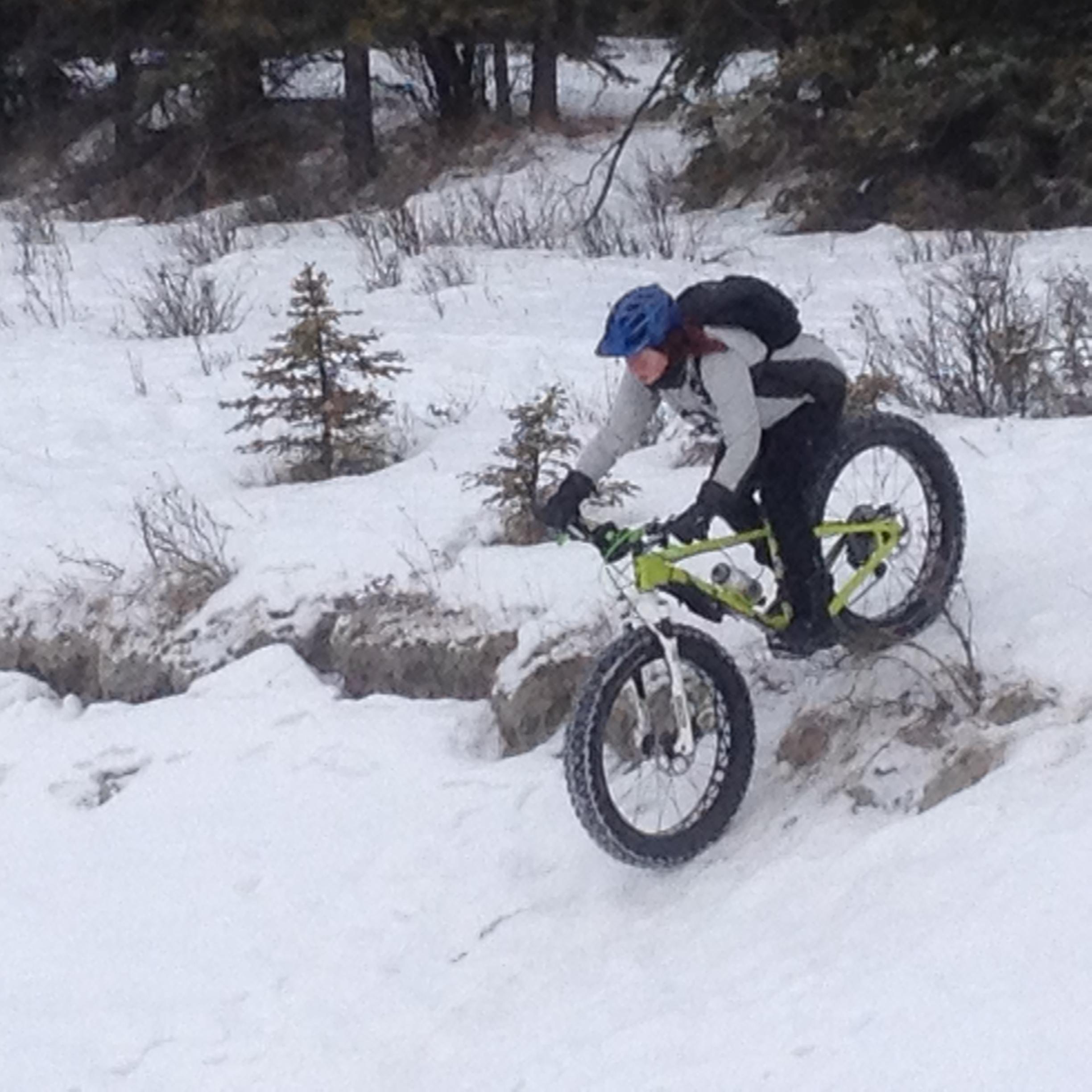 Specialized Fatboy Pro: A person riding a fat bike down a snowy slope, skillfully navigating through a winter landscape with sparse trees in the background. The rider is wearing a blue helmet and is dressed in winter attire, focused on maintaining balance as they descend.