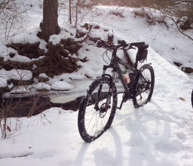 Mountain bike parked on a snowy trail beside a small stream, surrounded by trees. The bike has snow covering its tires and frame, indicating recent use in winter conditions. Trails seperated by streets mountain bike trail.