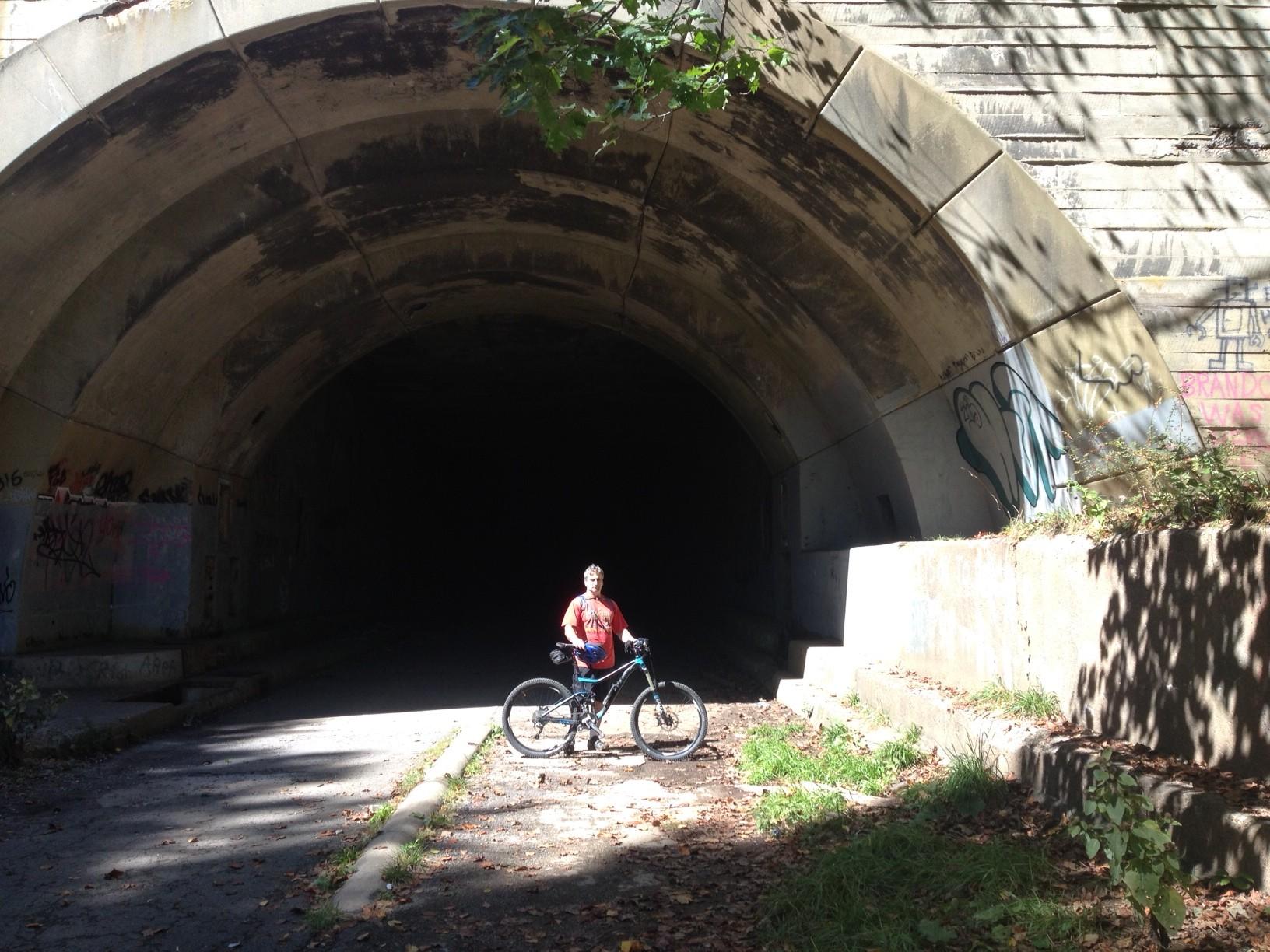 A person standing next to a bicycle at the entrance of a dark tunnel, surrounded by graffiti and natural foliage, with sunlight illuminating the area in front. Pike 2 Bike mountain bike trail.