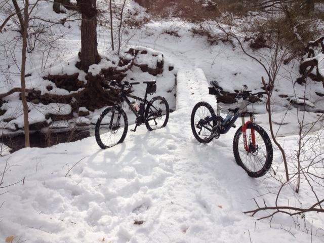 Two mountain bikes are positioned on a snowy path beside a small creek. The surrounding area features bare trees and snow-covered ground, creating a wintry outdoor scene. Sunshine is filtering through the trees, casting soft light on the bikes and the snow. Trails seperated by streets mountain bike trail.