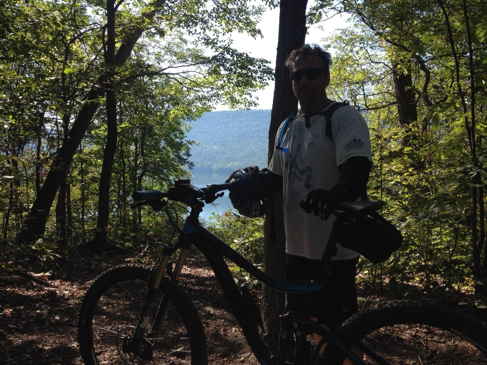 A person stands next to a mountain bike in a wooded area, wearing outdoor gear and sunglasses. The background features trees and a view of a calm lake surrounded by hills under a clear sky. The scene conveys a sense of adventure and nature. Allegrippis Trails mountain bike trail.