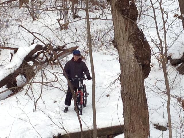 A person wearing a helmet and winter clothing stands next to a bicycle on a snowy path surrounded by bare trees and fallen branches. Trails seperated by streets mountain bike trail.