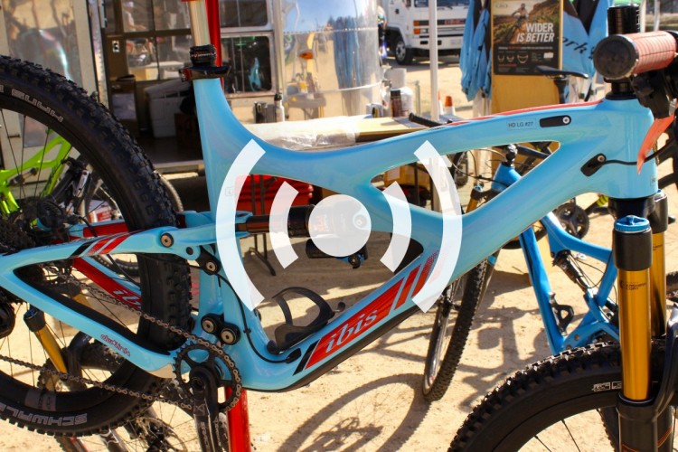 Alt text: Close-up view of a blue mountain bike frame featuring red and black accents, with a visible suspension system and tires in the background.