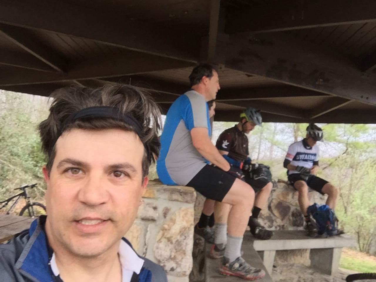 A group of four cyclists taking a break under a wooden shelter in a wooded area. One man is in the foreground, wearing a headband and a jacket, while three others sit on a stone bench behind him, dressed in cycling gear. The surrounding landscape shows trees and hints of greenery in the background. Sheltowee Trace - Laurel Lake Trail mountain bike trail.