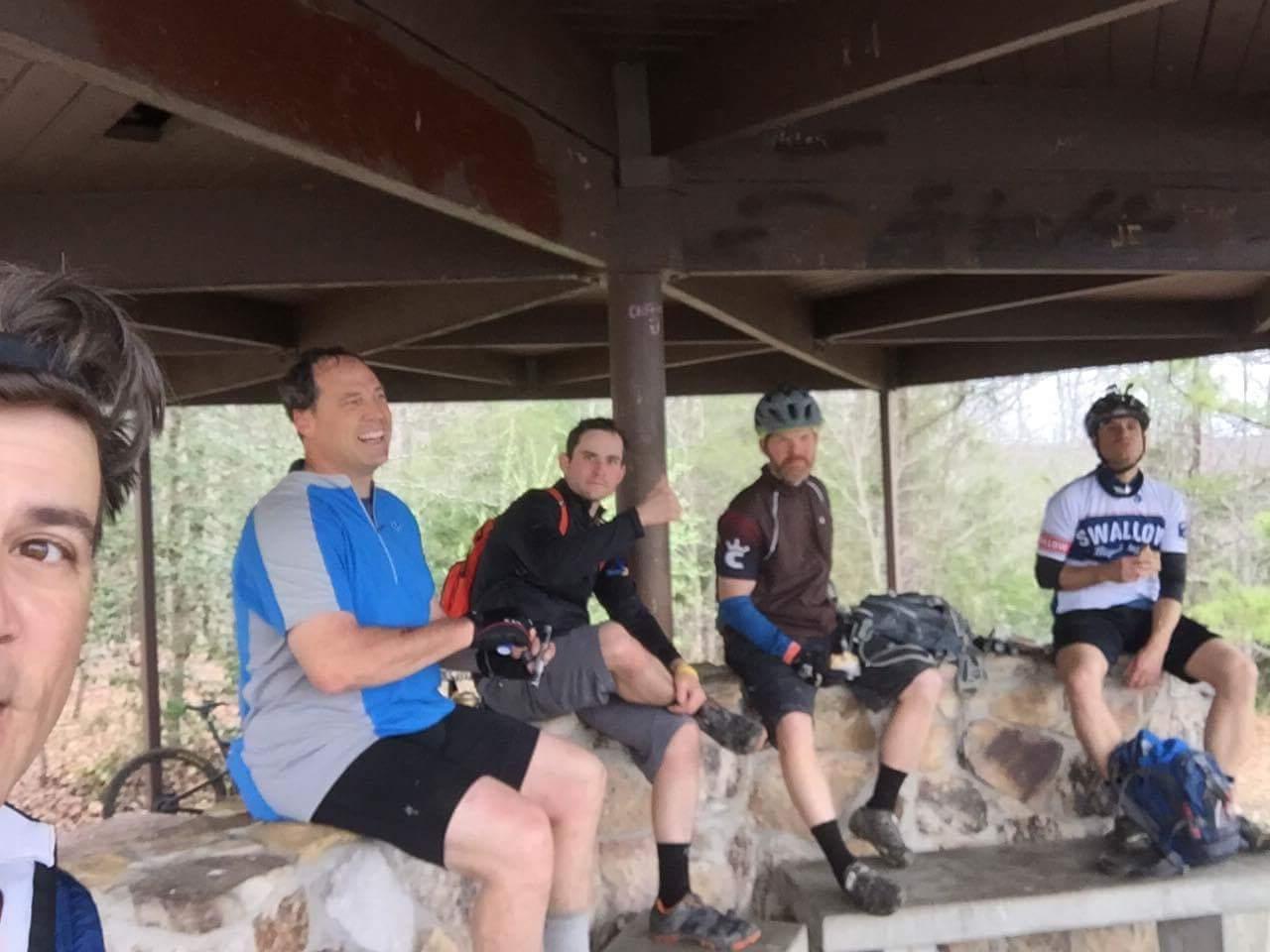 A group of five men sitting together on a stone bench under a shelter in a forested area, all dressed in cycling gear. They appear to be taking a break from biking, enjoying each other's company, and smiling. Sheltowee Trace - Laurel Lake Trail mountain bike trail.