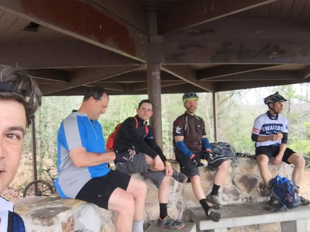 A group of five mountain bikers sitting on a stone bench under a shelter in a wooded area. They are wearing cycling gear, including helmets and padded shorts, and appear to be taking a break. Some are checking their gear, while others are engaged in conversation. Sheltowee Trace - Laurel Lake Trail mountain bike trail.