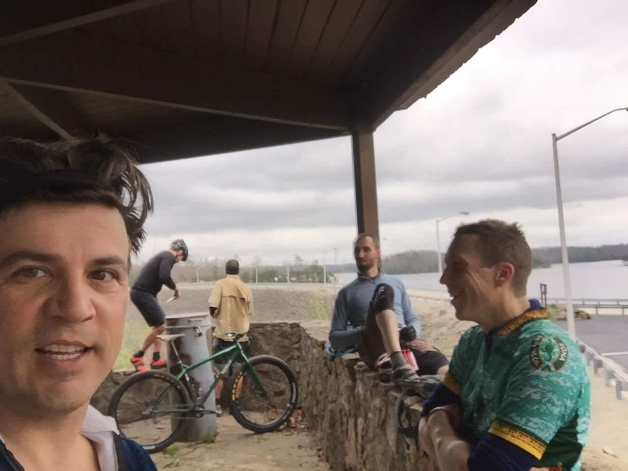 A group of four individuals, including one in the foreground taking a selfie, gather under a shelter near a body of water. Two people are seated on a stone wall, while another stands nearby wearing a helmet and preparing a bike. The scene is set against a cloudy sky, with trees visible in the background. Sheltowee Trace - Laurel Lake Trail mountain bike trail.