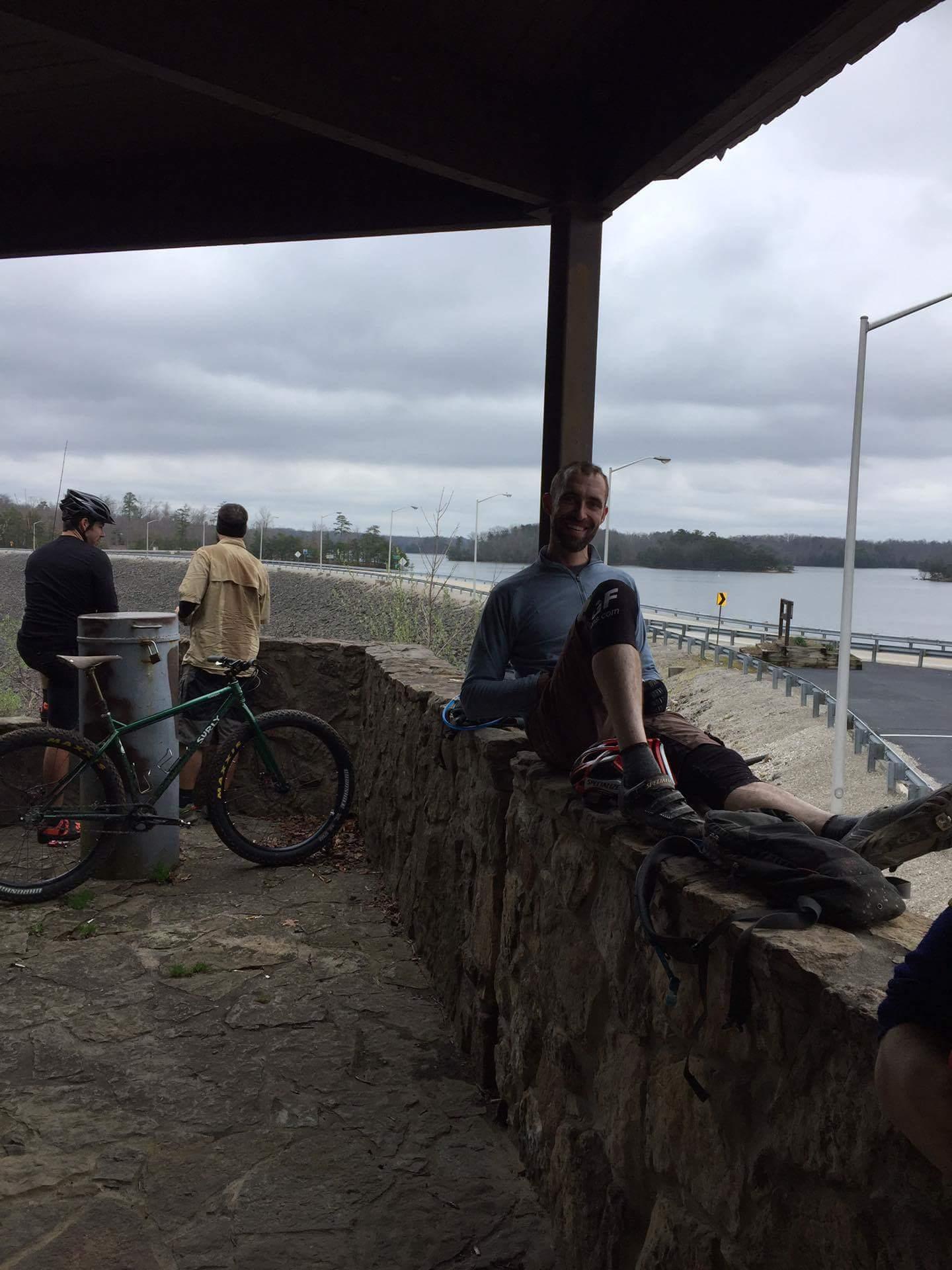 A group of people resting on a stone structure with a view of a lake and cloudy sky. One person is sitting on the wall, smiling, while others are standing near their bicycles. The setting appears to be an outdoor recreational area. Sheltowee Trace - Laurel Lake Trail mountain bike trail.
