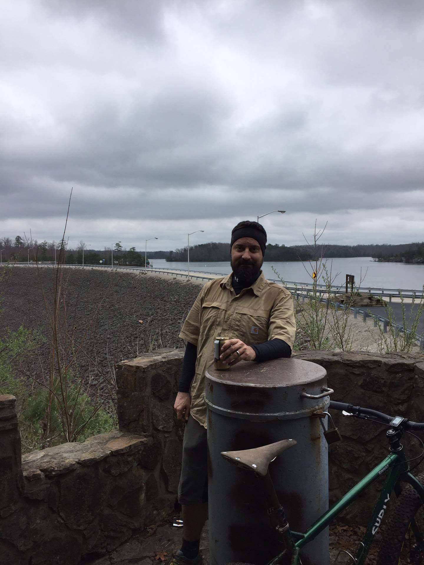 A person wearing a tan short-sleeved shirt and a black beanie stands next to a metal trash can, holding a can drink. In the background, there is a cloudy sky, a body of water, and trees, with a road visible. A green bicycle is positioned nearby. Sheltowee Trace - Laurel Lake Trail mountain bike trail.