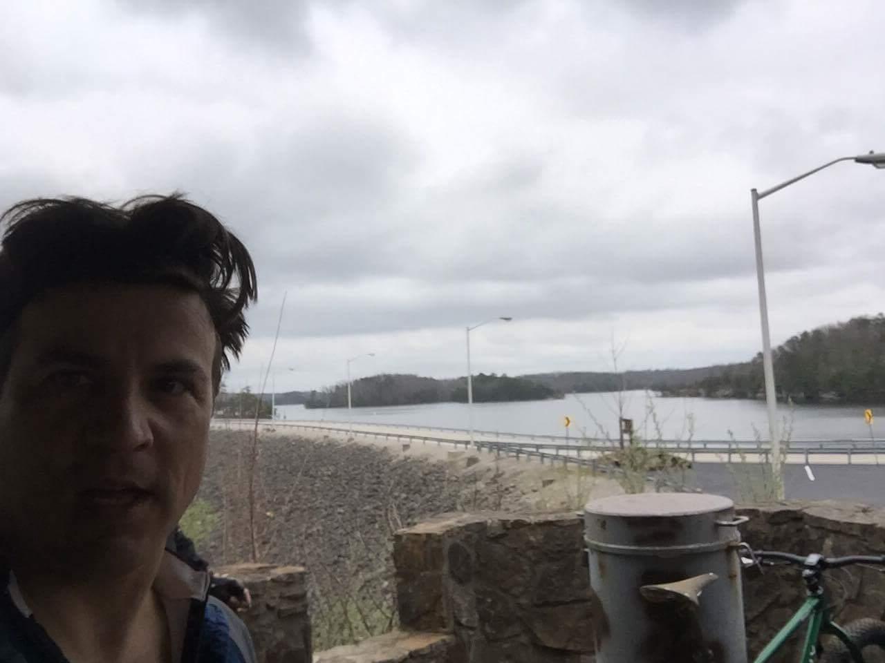 Selfie of a person with tousled hair, standing near a stone railing. In the background, a lake is visible with a road running alongside it, surrounded by trees under an overcast sky. A bicycle is leaning against the stone railing next to a trash can. Sheltowee Trace - Laurel Lake Trail mountain bike trail.