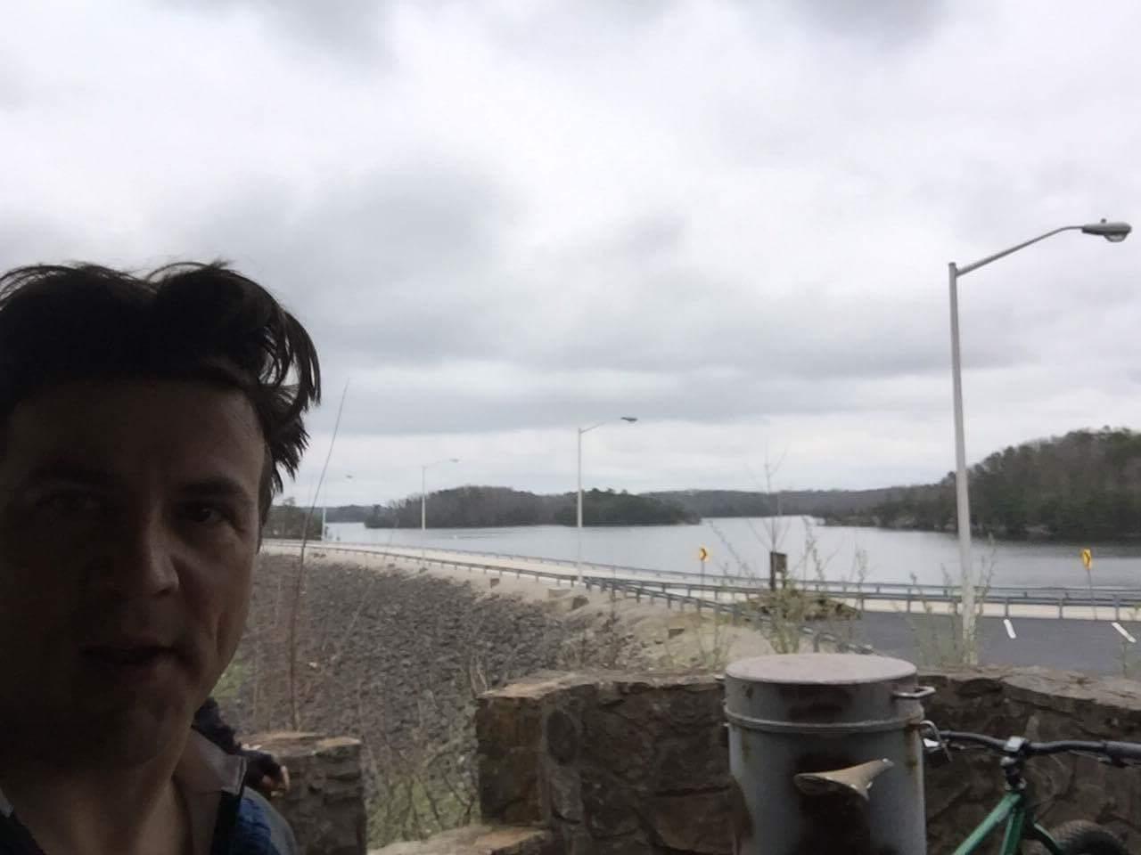 A person takes a selfie near a stone wall overlooking a river with trees in the background on a cloudy day. A road runs alongside the river, and a bicycle is parked nearby. Sheltowee Trace - Laurel Lake Trail mountain bike trail.