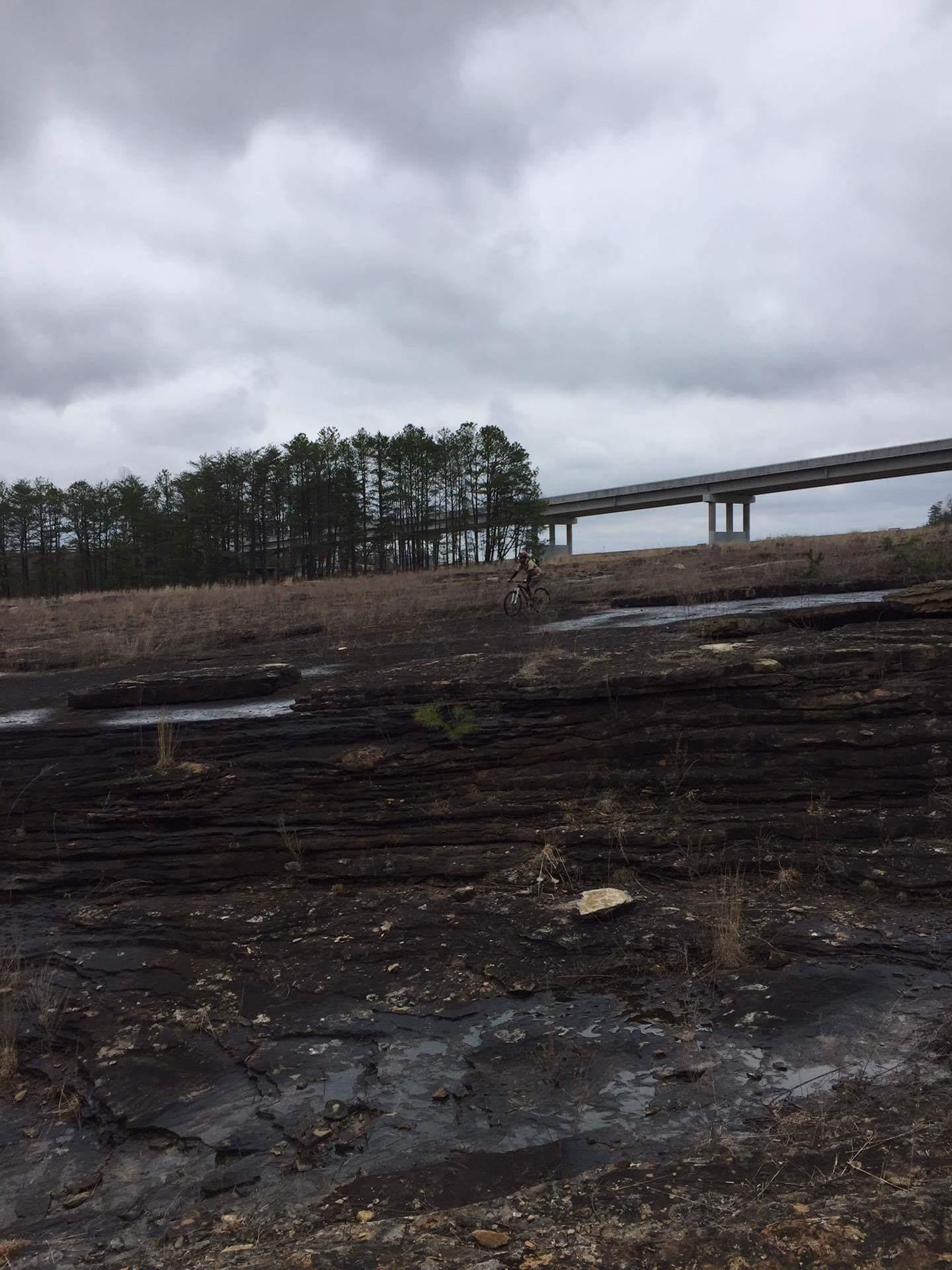 A barren landscape with rocky terrain and patches of grass, under a cloudy sky. In the background, there is a bridge with tall pillars crossing over the area, and a few tall pine trees line the horizon. A solitary mountain bike is positioned near the rocky area. Sheltowee Trace - Laurel Lake Trail mountain bike trail.