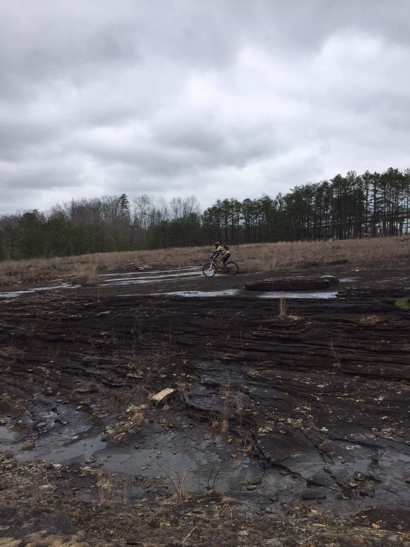 A person riding a mountain bike on a rocky, uneven terrain under a cloudy sky, with sparse vegetation and a line of trees in the background. Sheltowee Trace - Laurel Lake Trail mountain bike trail.