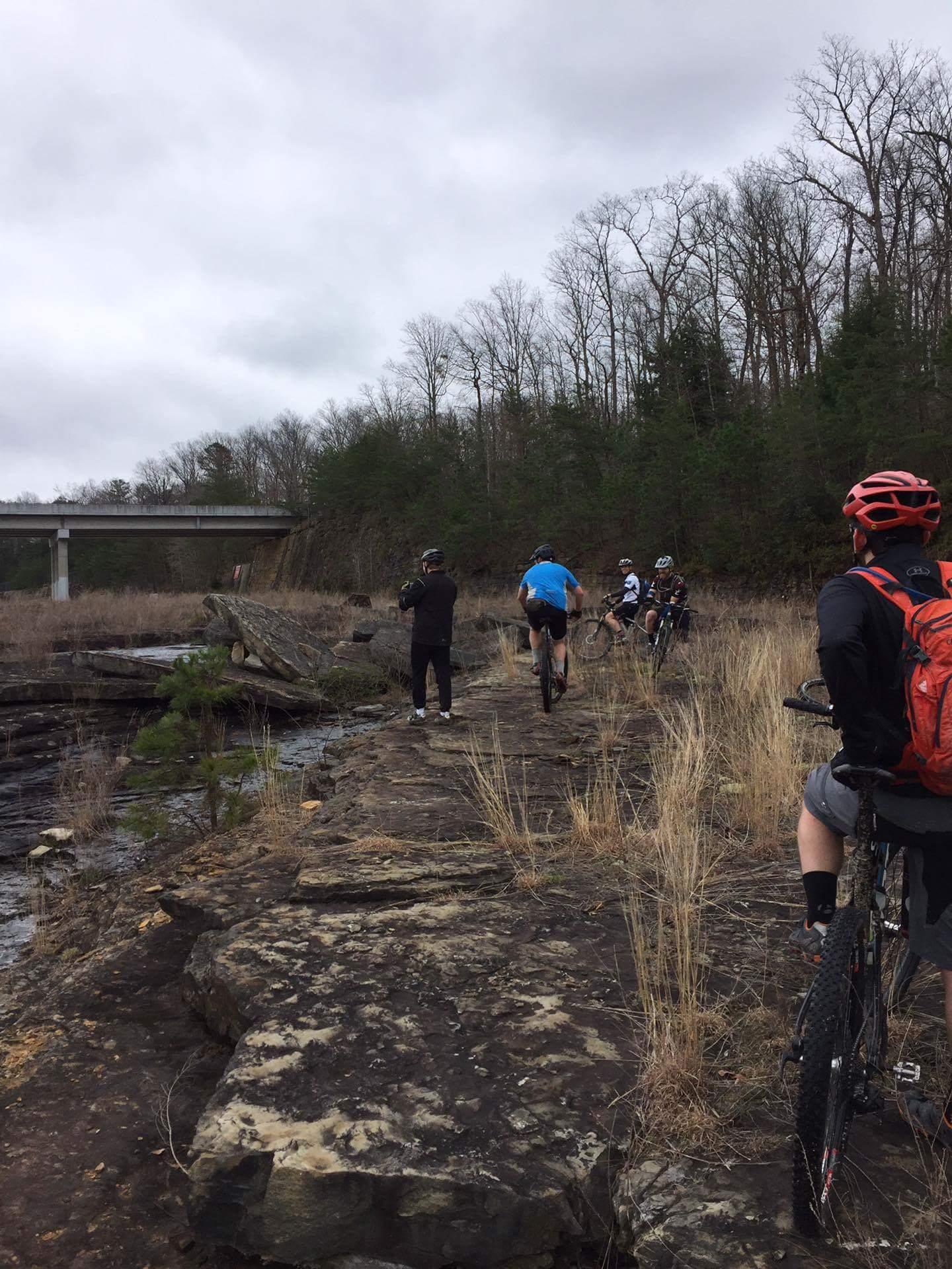 A group of mountain bikers exploring a rocky trail near a waterway, with a bridge visible in the background. The setting is surrounded by sparse trees and tall grass under a cloudy sky. Sheltowee Trace - Laurel Lake Trail mountain bike trail.
