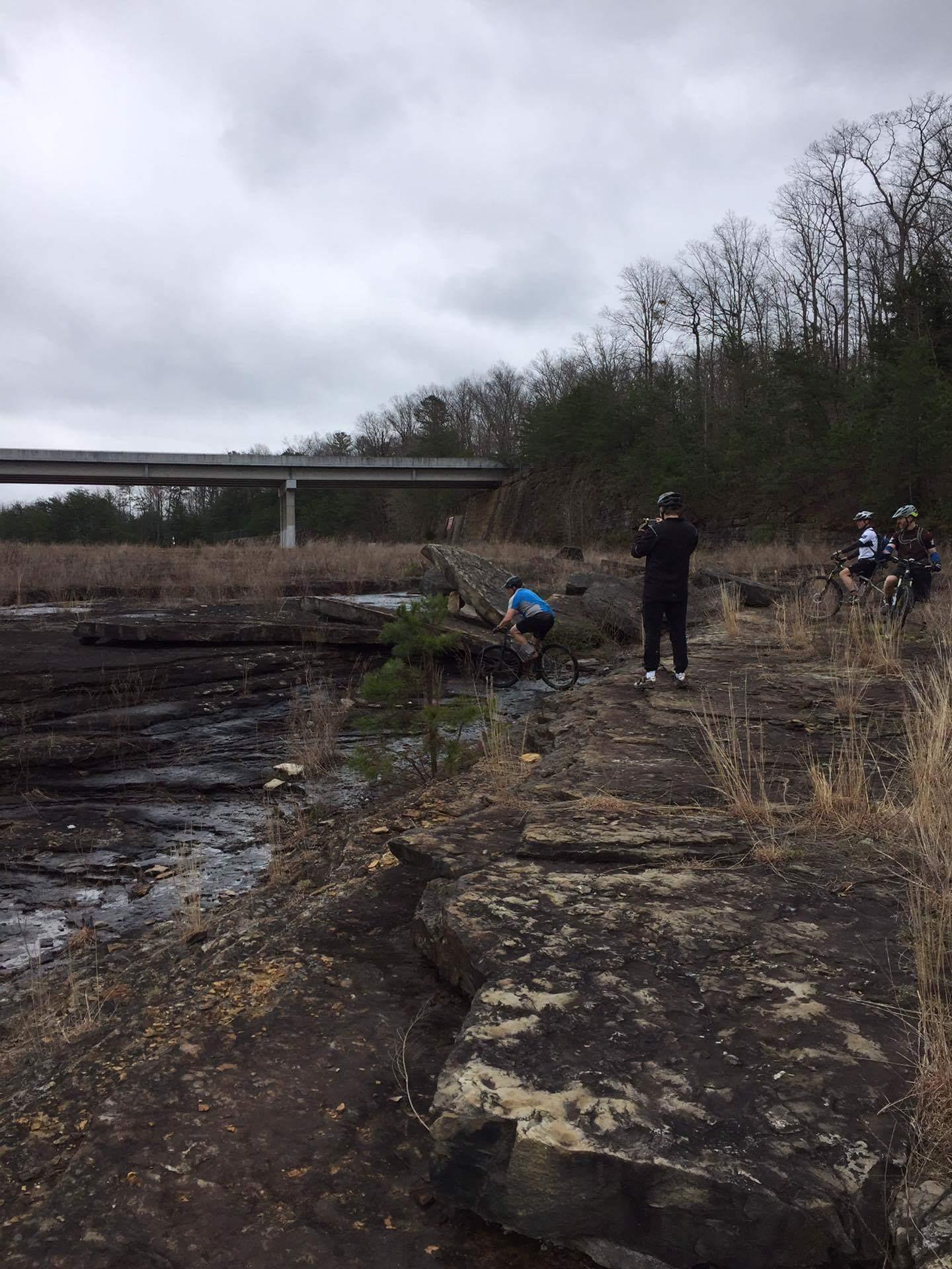 A group of mountain bikers and a photographer are gathered near a rocky area by a riverbank, with a bridge visible in the background. The sky is overcast, and the landscape features sparse vegetation and bare trees, suggesting a cool, possibly early spring environment. One biker is adjusting their bike while others stand nearby, enjoying the scenery. Sheltowee Trace - Laurel Lake Trail mountain bike trail.