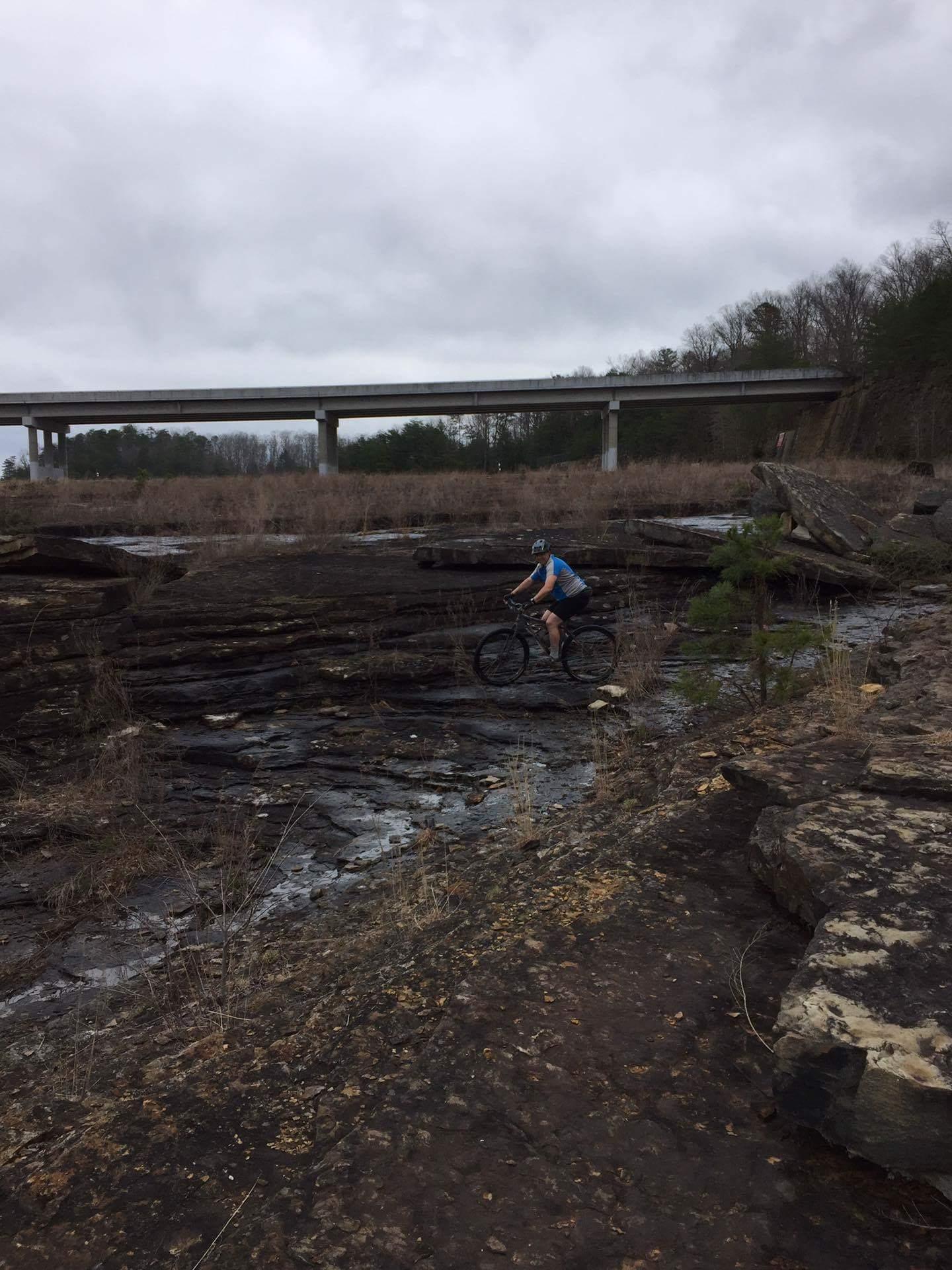 A cyclist riding along a rocky terrain with sparse grass, under a cloudy sky. In the background, a concrete bridge crosses over the landscape, surrounded by trees. Sheltowee Trace - Laurel Lake Trail mountain bike trail.