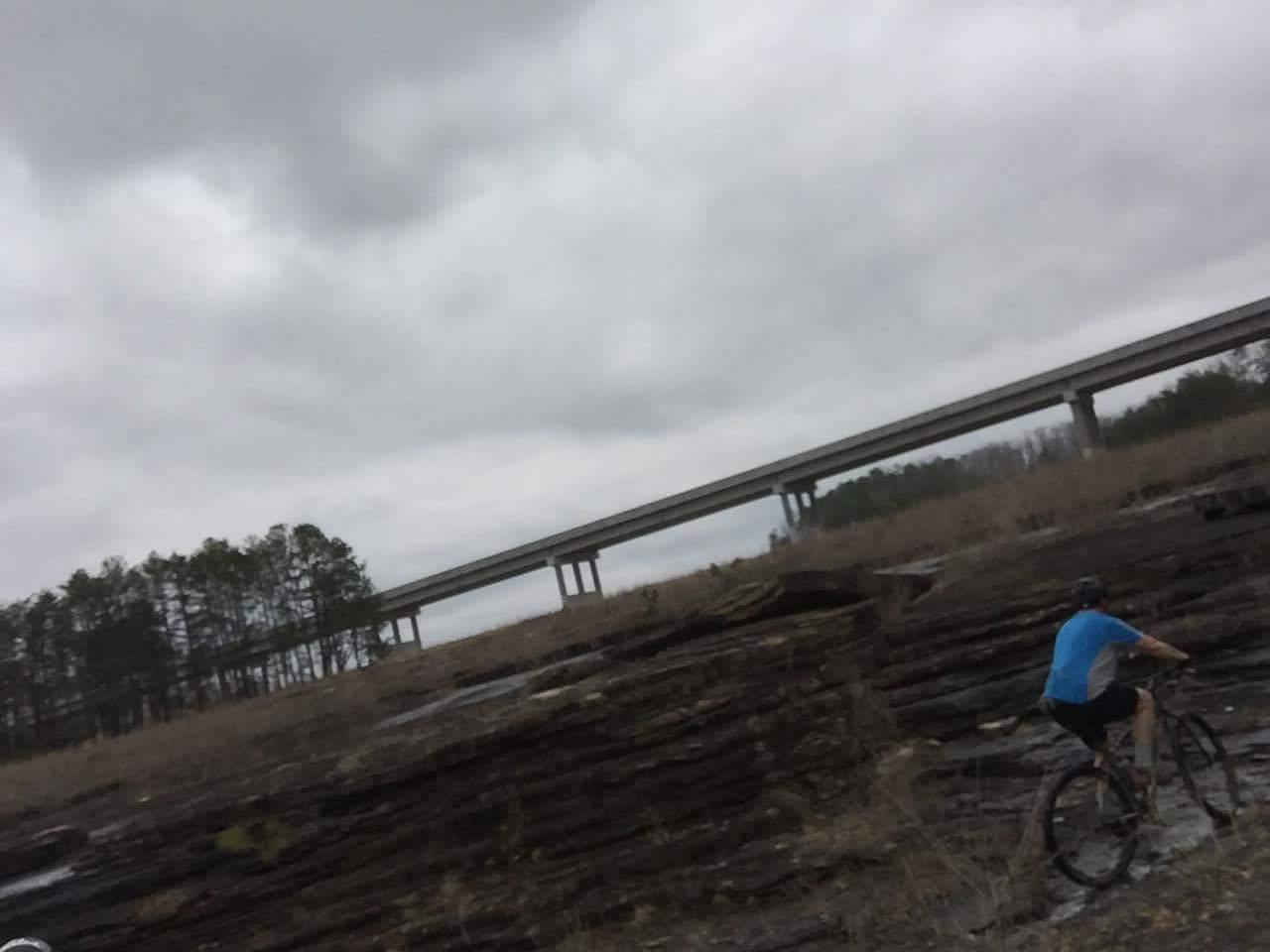 A mountain biker riding on rocky terrain beneath a cloudy sky, with a highway overpass in the background. The landscape features sparse vegetation and a mix of dried grass and rocks. Sheltowee Trace - Laurel Lake Trail mountain bike trail.