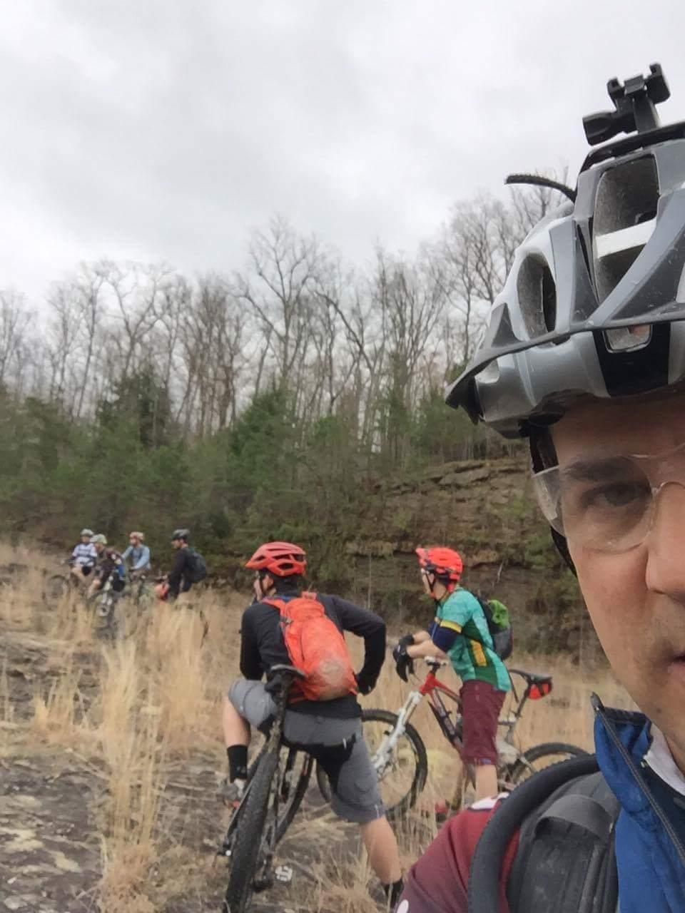 A group of mountain bikers pauses on a rocky trail surrounded by sparse vegetation and trees under a cloudy sky. The foreground features a person wearing a helmet and glasses, taking a selfie while several cyclists are visible in the background, some wearing bright helmets and backpacks. Sheltowee Trace - Laurel Lake Trail mountain bike trail.