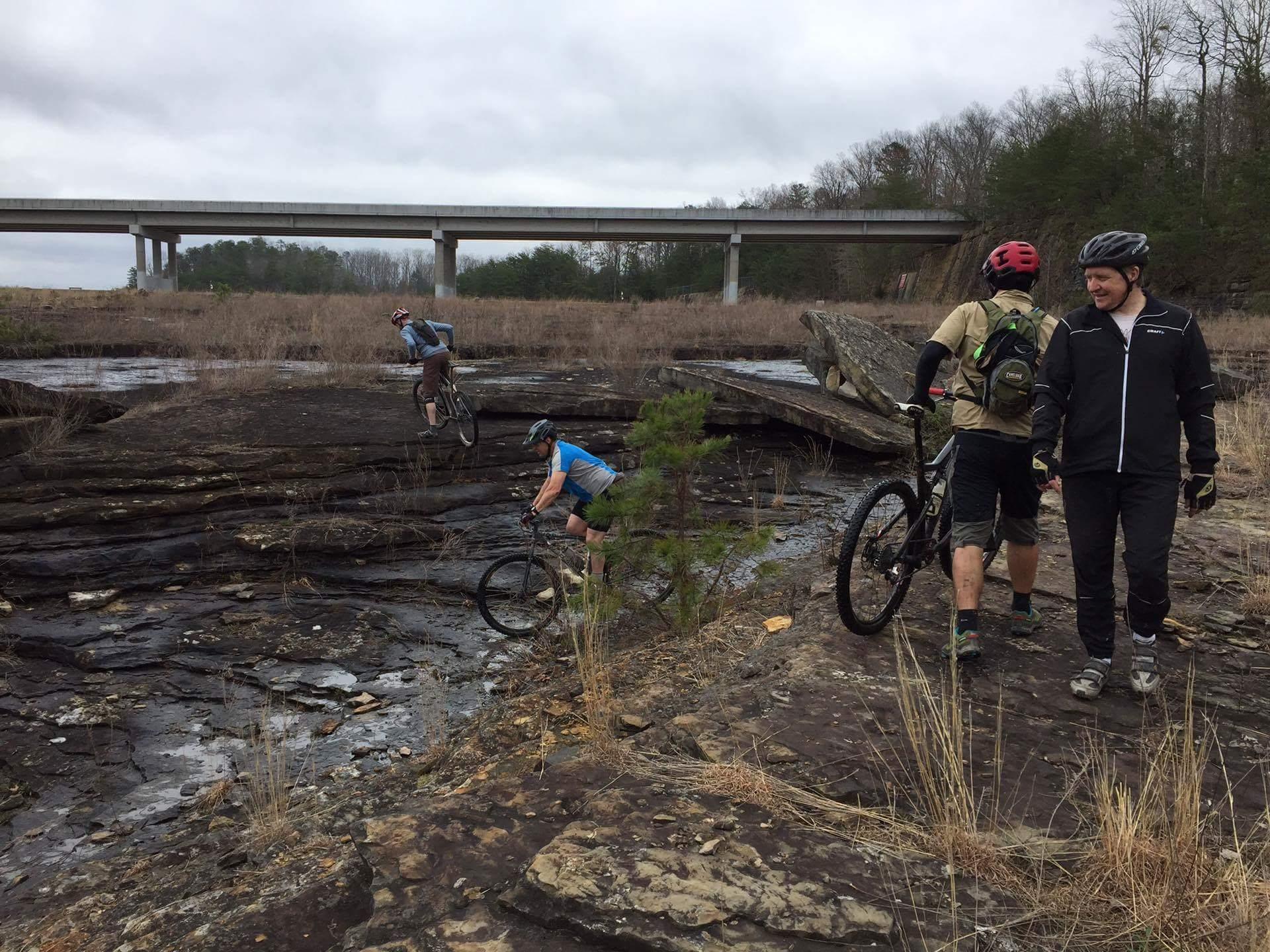 A group of mountain bikers navigating rocky terrain near a water source, with a bridge visible in the background. One biker is descending a rocky slope, while another is walking beside their bike, and a third biker is preparing to ride. The area features sparse vegetation and a cloudy sky. Sheltowee Trace - Laurel Lake Trail mountain bike trail.
