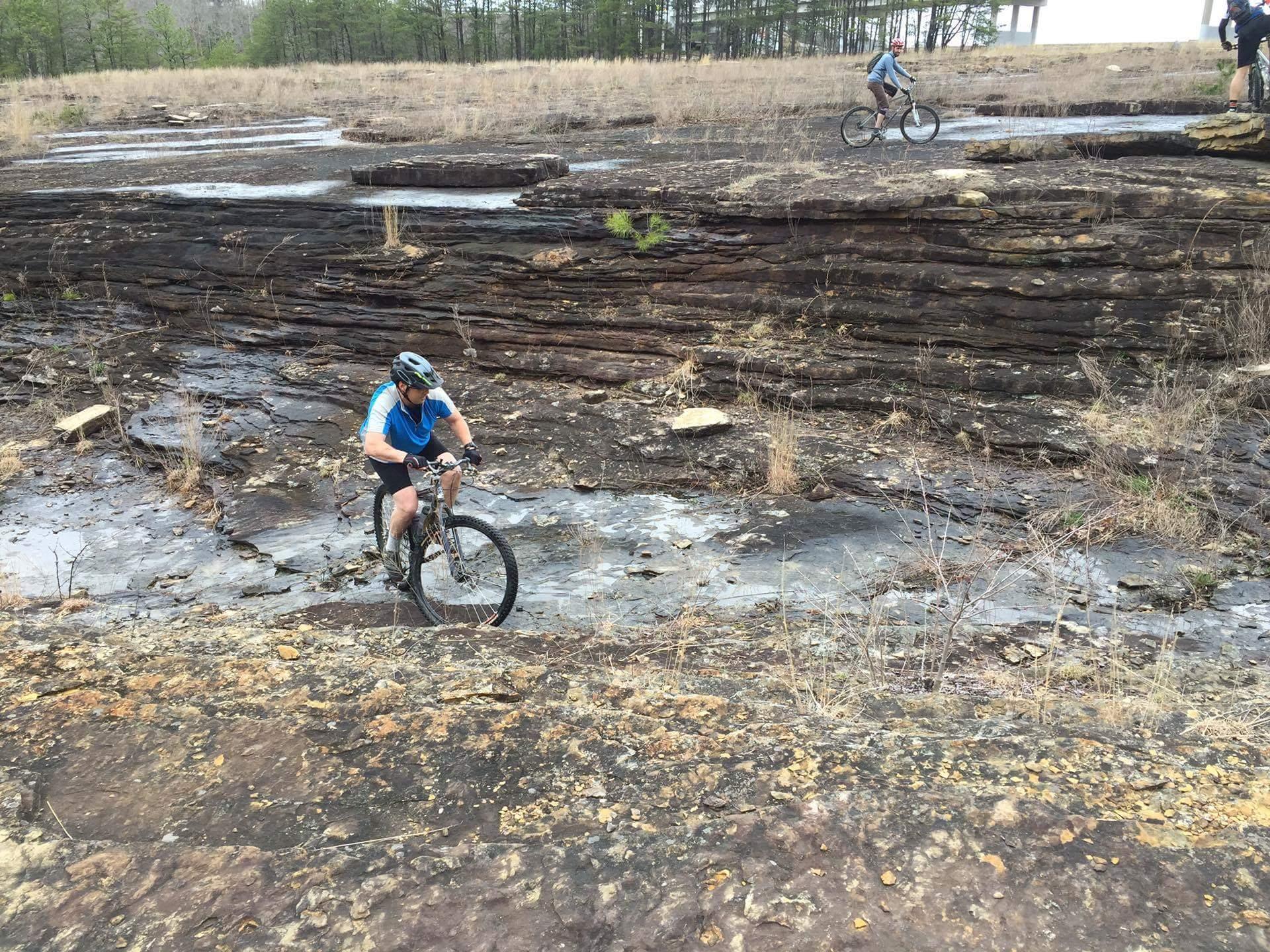 A mountain biker navigating a rocky terrain with layered stone formations and sparse vegetation. In the background, another cyclist is riding on a similar path, showcasing an outdoor cycling scene. Sheltowee Trace - Laurel Lake Trail mountain bike trail.