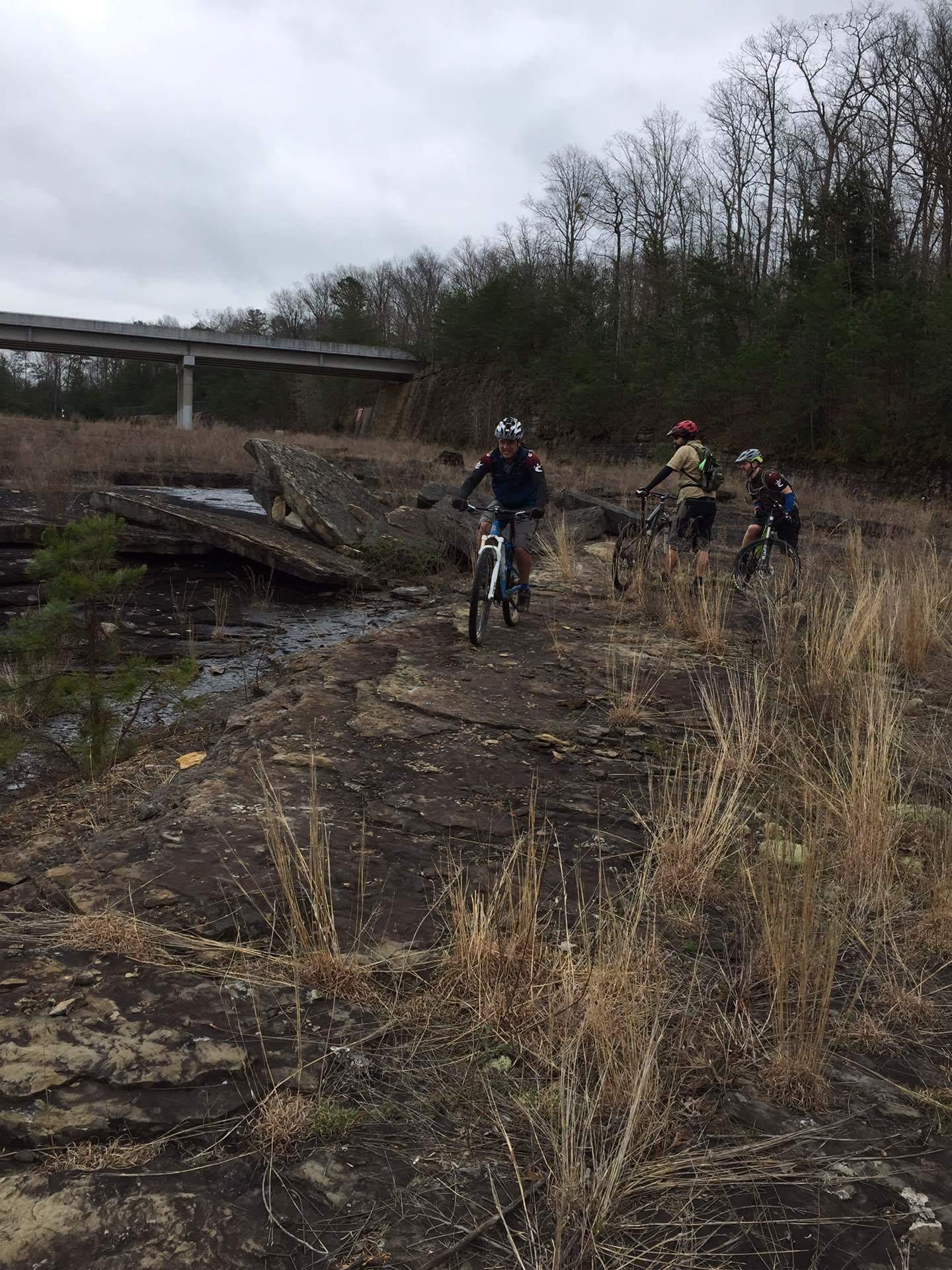 Three mountain bikers navigate a rocky, grassy terrain near a creek. In the background, a highway overpass is visible, surrounded by trees. The sky is overcast, suggesting a cloudy day. Sheltowee Trace - Laurel Lake Trail mountain bike trail.