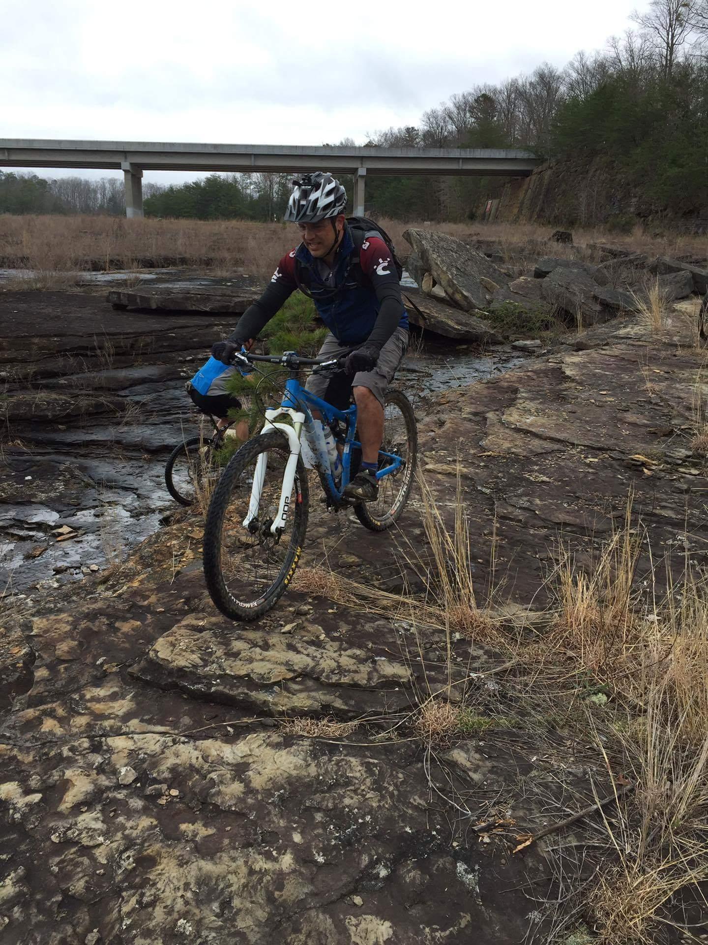 Mountain biker navigating rocky terrain near a river, with dry grass in the foreground and a bridge visible in the background. The cyclist is dressed in a helmet and sports gear, riding a blue bike over the uneven surface. Sheltowee Trace - Laurel Lake Trail mountain bike trail.