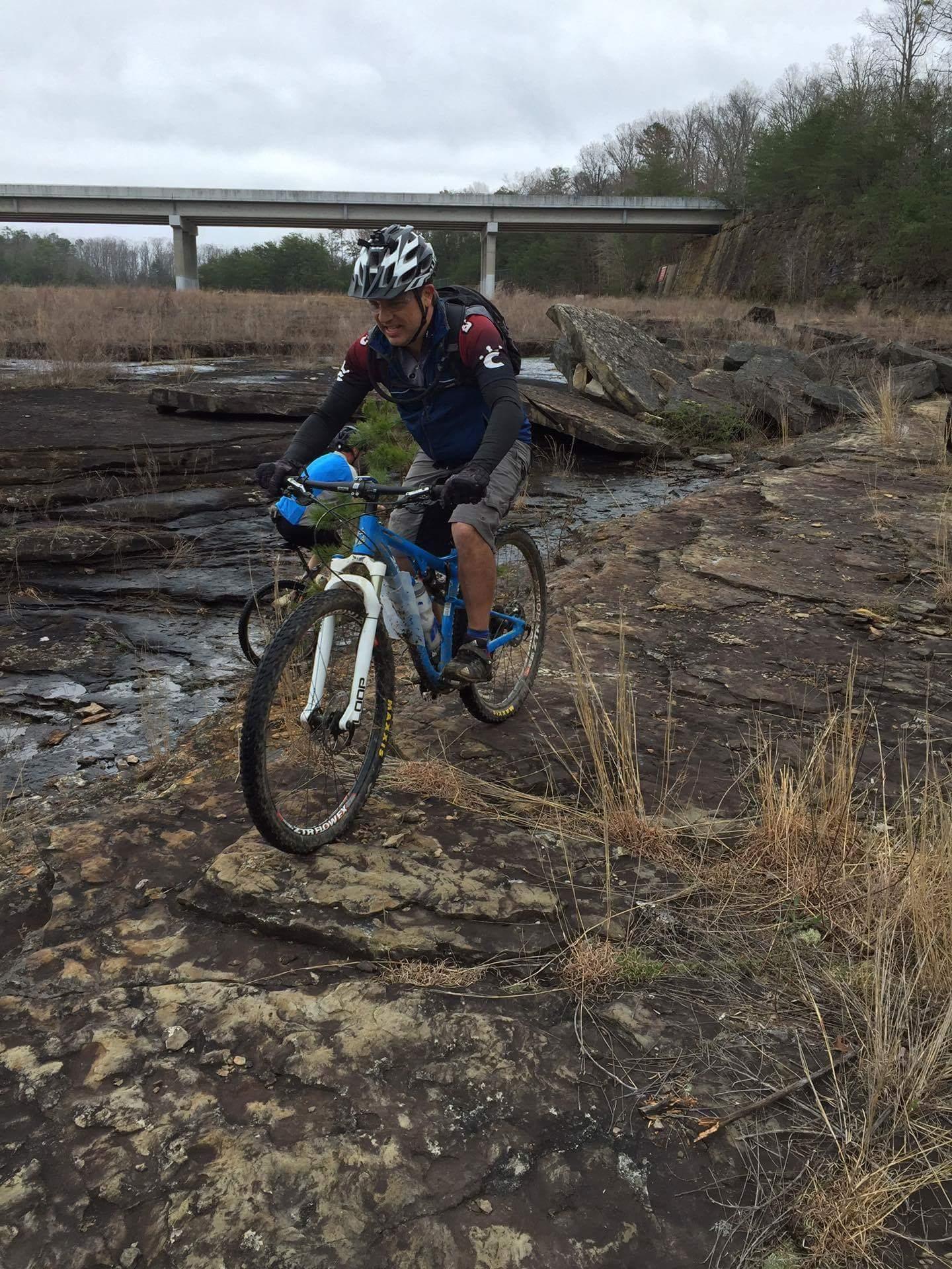A mountain biker traverses rocky terrain near a river, with dry grasses and scattered stones surrounding him. A bridge spans the background under a cloudy sky. The cyclist wears a helmet and cycling attire, focused on navigating the challenging landscape on a blue mountain bike. Sheltowee Trace - Laurel Lake Trail mountain bike trail.