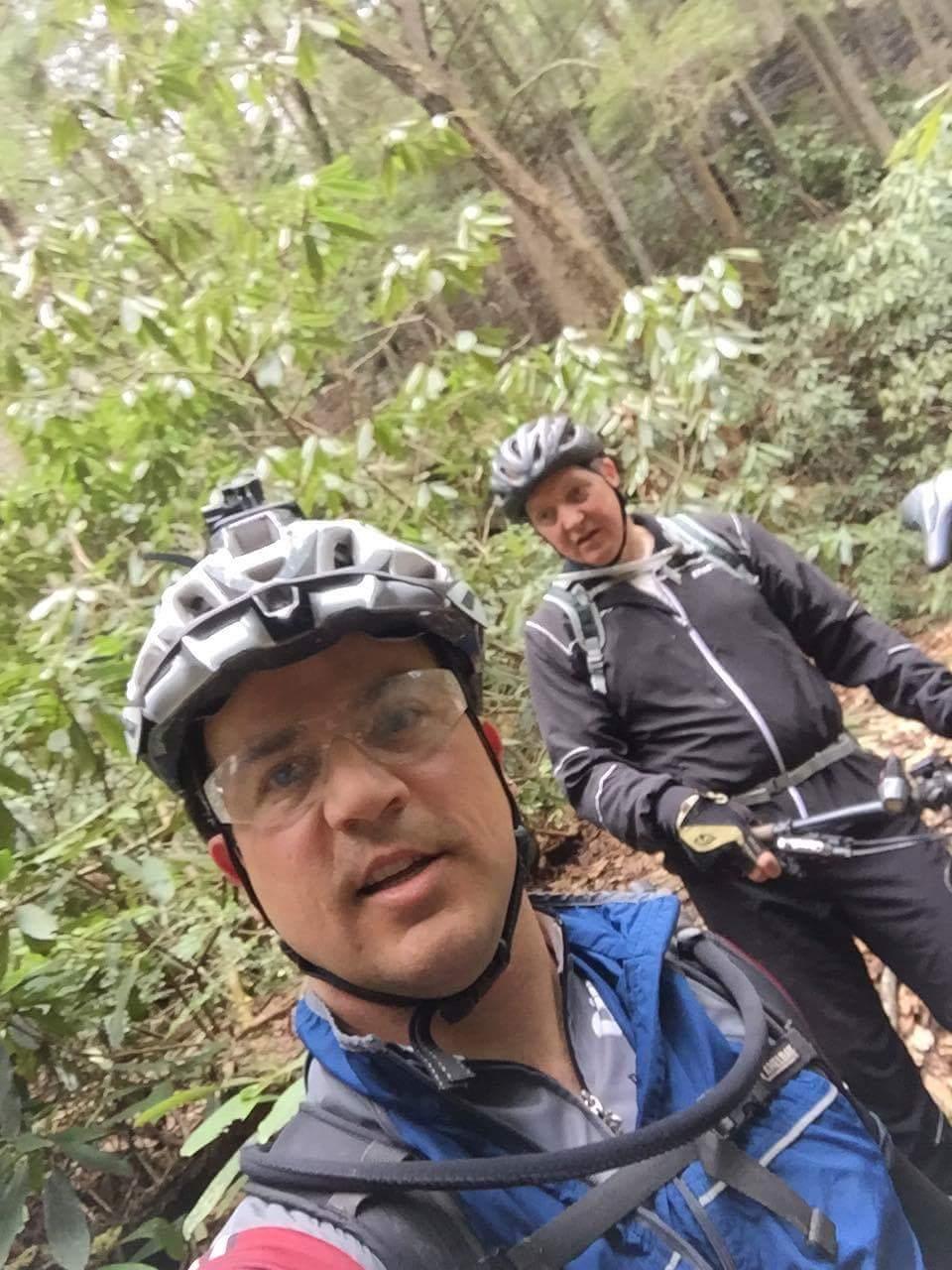 Two men wearing helmets and wearing cycling gear are taking a selfie while mountain biking in a lush green forest. The dense foliage surrounds them, creating a natural backdrop for their outdoor adventure. One man is smiling at the camera, while the other looks focused and slightly turned away from the camera. Both appear to be enjoying their experience in the great outdoors. Sheltowee Trace - Laurel Lake Trail mountain bike trail.