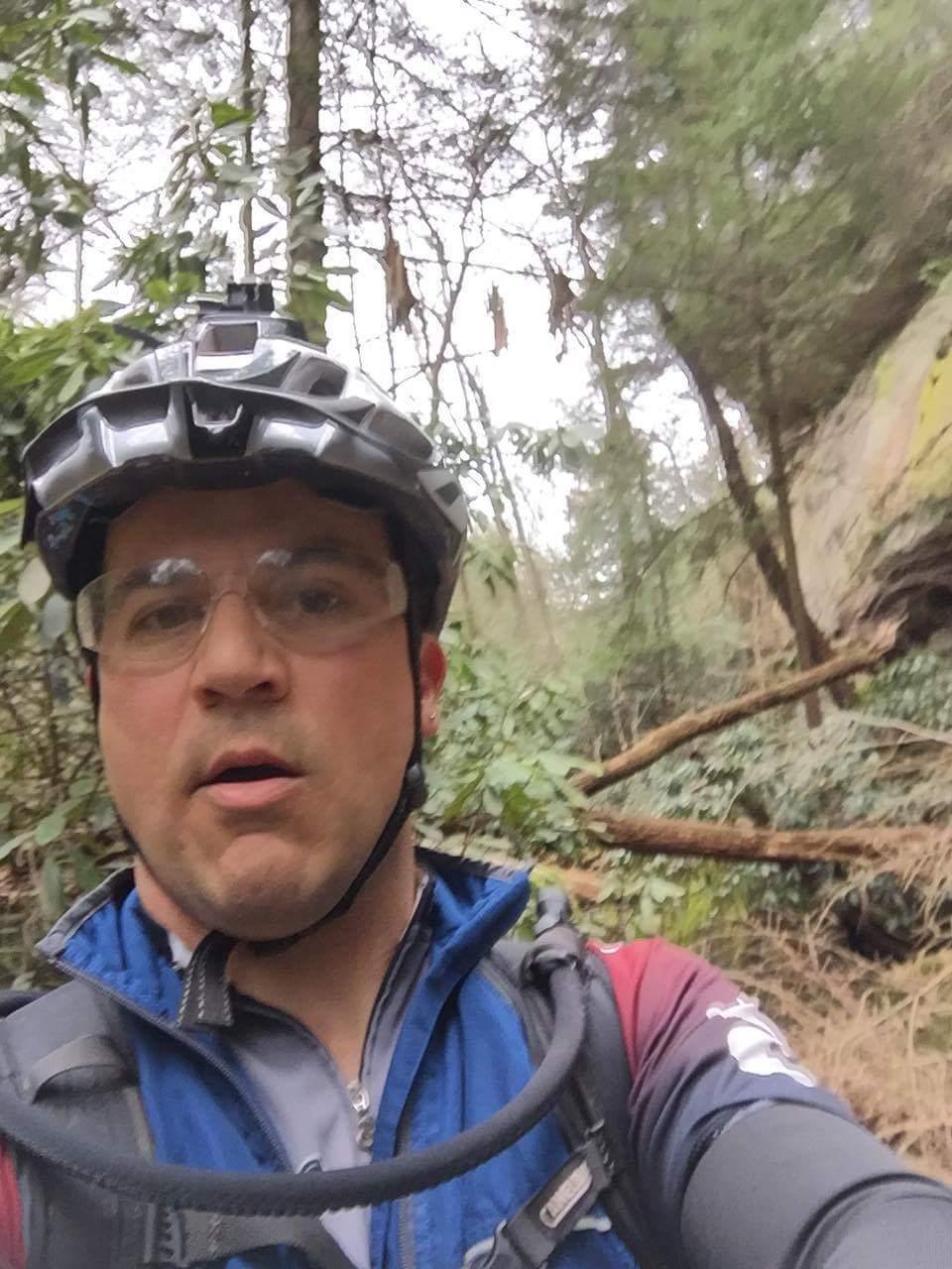 A person wearing a helmet and glasses takes a selfie while biking on a wooded trail surrounded by trees and greenery. The landscape includes fallen branches and natural vegetation, indicating an outdoor adventure. The atmosphere appears overcast and serene. Sheltowee Trace - Laurel Lake Trail mountain bike trail.