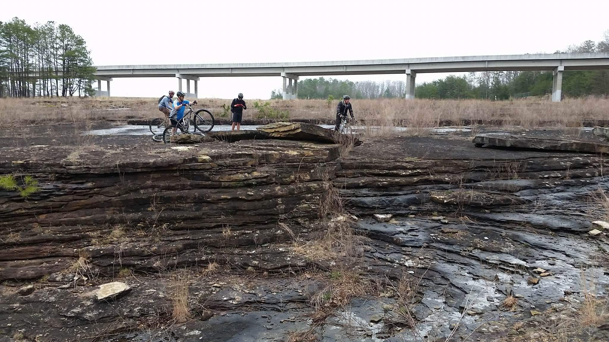 Three mountain bikers are positioned on a rocky landscape with layered stone formations. In the background, a concrete overpass is visible, and sparse vegetation and dry grasses surround the area, suggesting an outdoor setting. The sky is overcast, indicating a cloudy day. Sheltowee Trace - Laurel Lake Trail mountain bike trail.