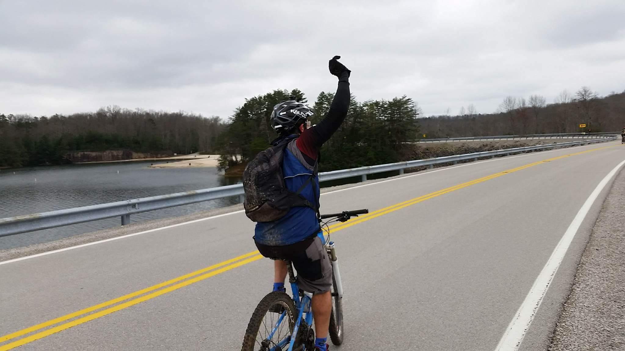 A cyclist wearing a helmet and a blue vest raises one hand while riding on a road near a calm body of water, surrounded by trees and a cloudy sky. The cyclist's bike is partially muddy, indicating recent use on trails. Sheltowee Trace - Laurel Lake Trail mountain bike trail.