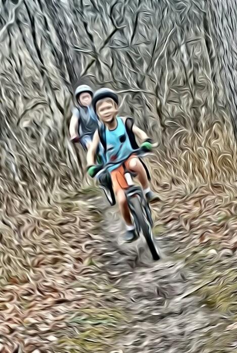 Two children riding bicycles on a wooded trail, with one child in the foreground and another slightly blurred in the background. The scene is characterized by a soft, artistic effect that adds a dreamy quality to the forest setting. Versailles State Park mountain bike trail.