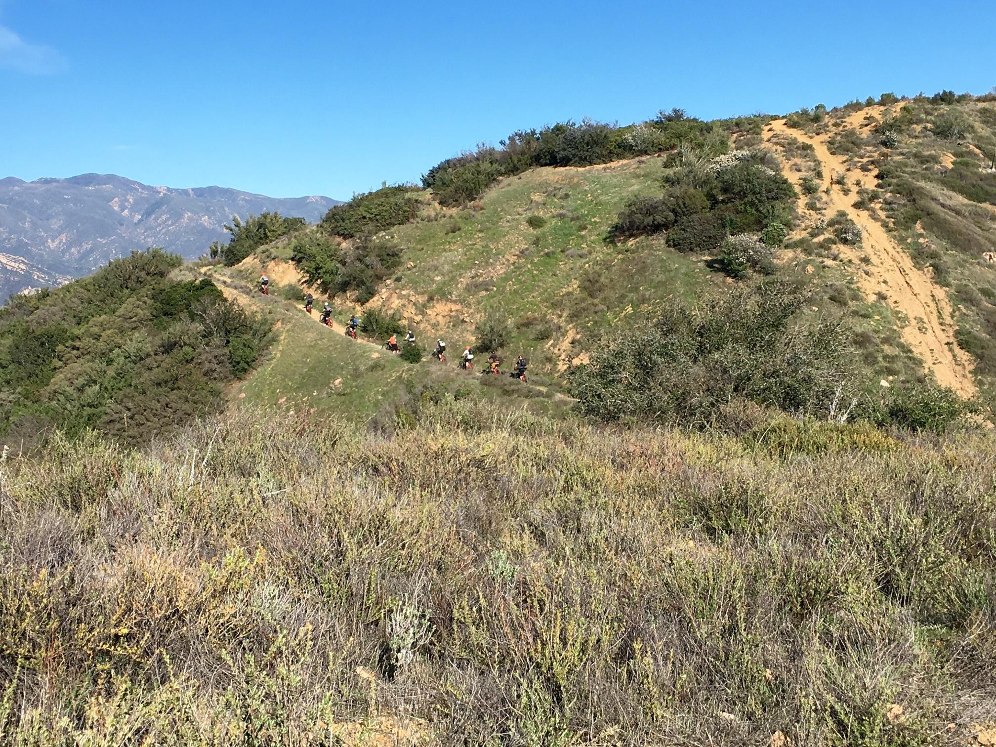 A scenic view of a mountain trail with several mountain bikers riding along a winding path. The background features rolling hills and distant mountains under a clear blue sky, with greenery and shrubs lining the trail. Romero Road mountain bike trail.