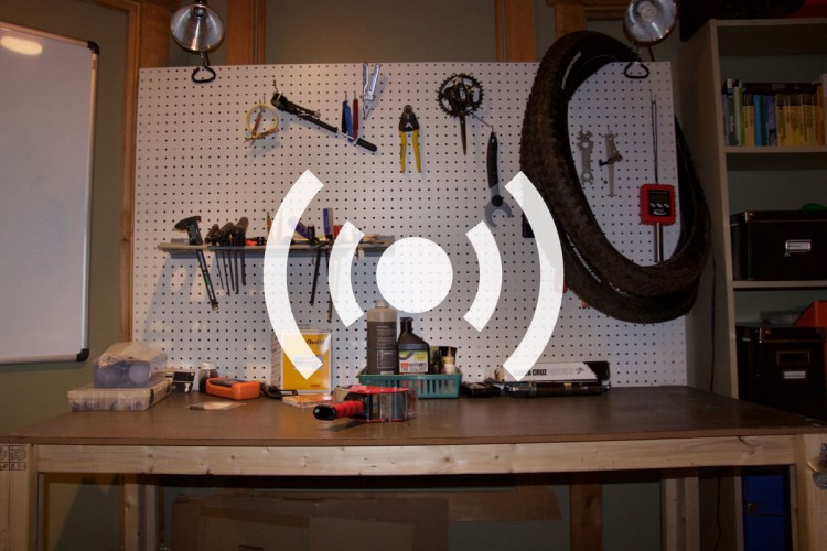 A well-organized workshop with a perforated wall displaying various hand tools, including wrenches, pliers, and screwdrivers. A work table is beneath, featuring a few toolboxes, oil containers, and a flashlight, with a bicycle tire hung on the wall above. A whiteboard is visible on the left side of the image.