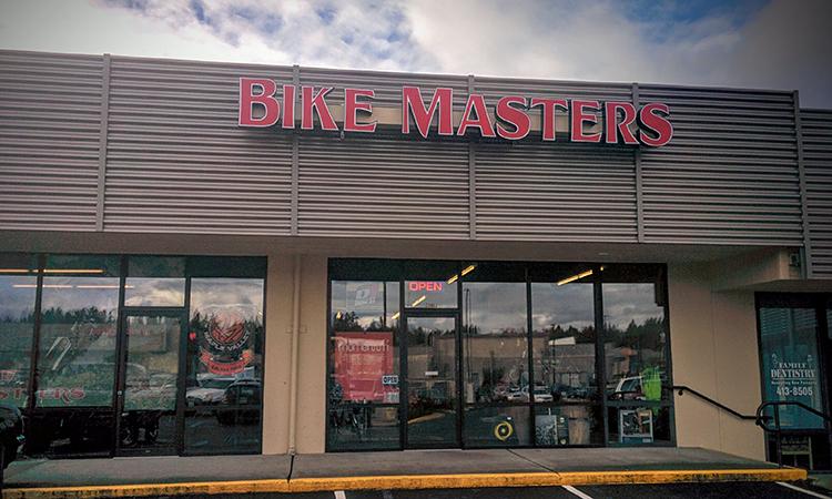 Exterior view of a store named "Bike Masters," featuring large red signage. The store has glass windows displaying various bike-related items and the word "OPEN" is visible in the window. The building is modern with a striped awning, and the sky above is partly cloudy.