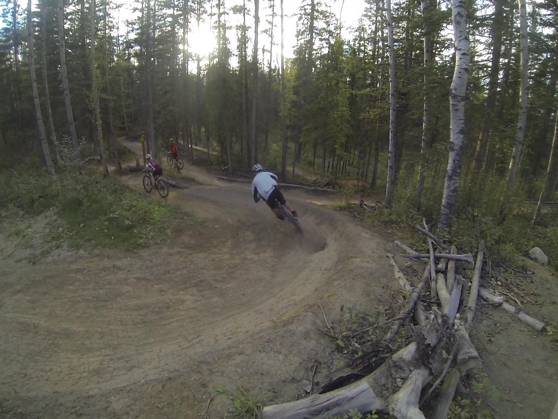 A group of mountain bikers maneuvering through a winding dirt trail in a forested area. The scene captures one rider leaning into a sharp turn while two other riders follow on a sunny day surrounded by tall trees. Hinton Skills Park mountain bike trail.