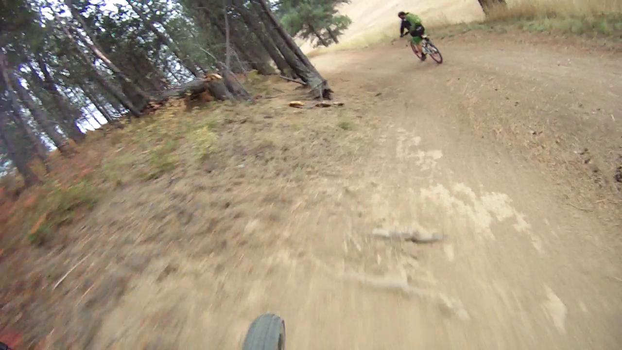 A mountain biker riding on a dirt trail through a forest, with trees lining the path on either side and a slight incline visible in the background. The perspective shows the front wheel of the bike and the rider ahead, capturing the motion and nature of the outdoor activity. Rattlesnake mountain bike trail.
