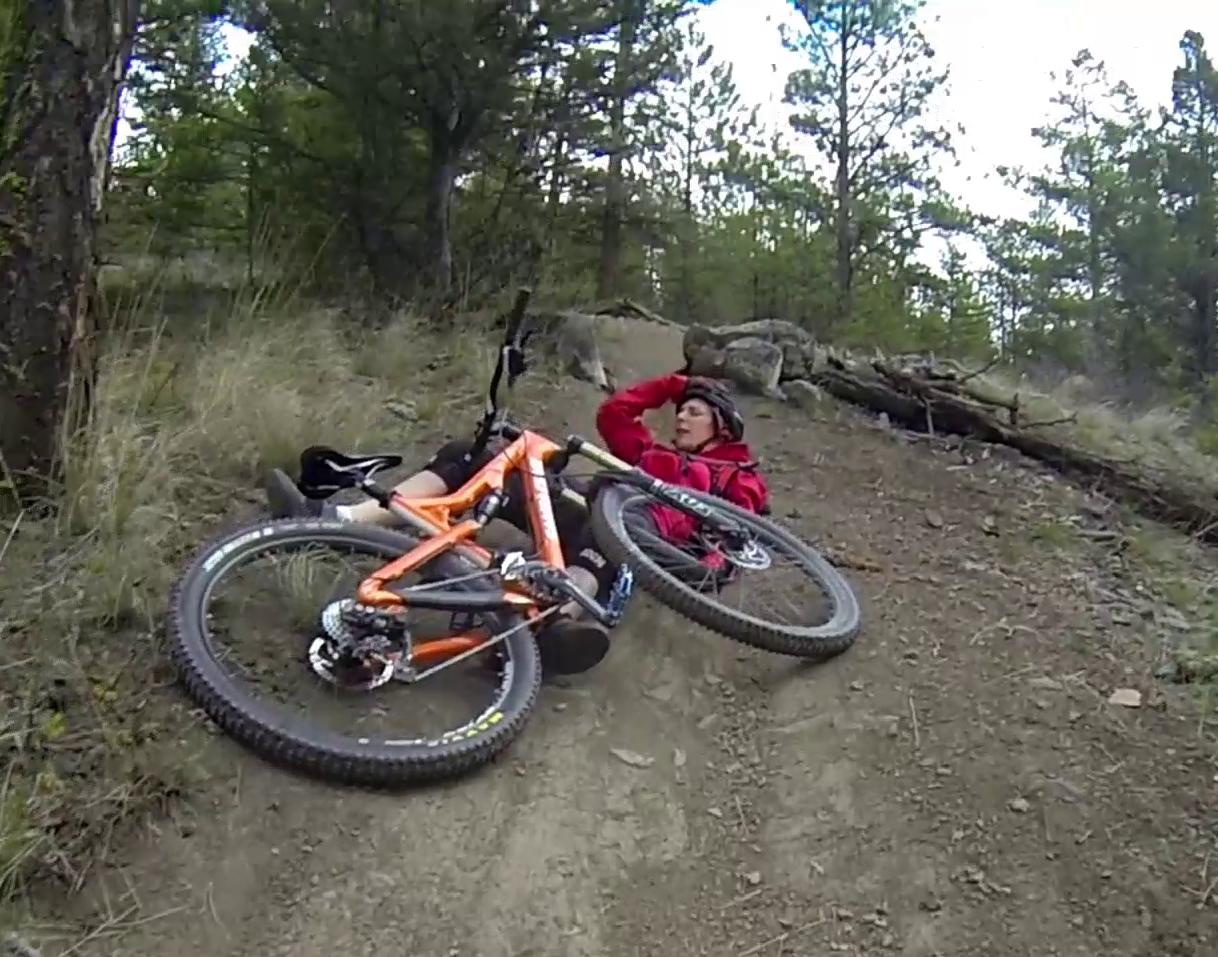 A mountain biker lying on the ground next to an orange bicycle, looking distressed, in a forested area with trees and dirt trails. Smith Creek mountain bike trail.
