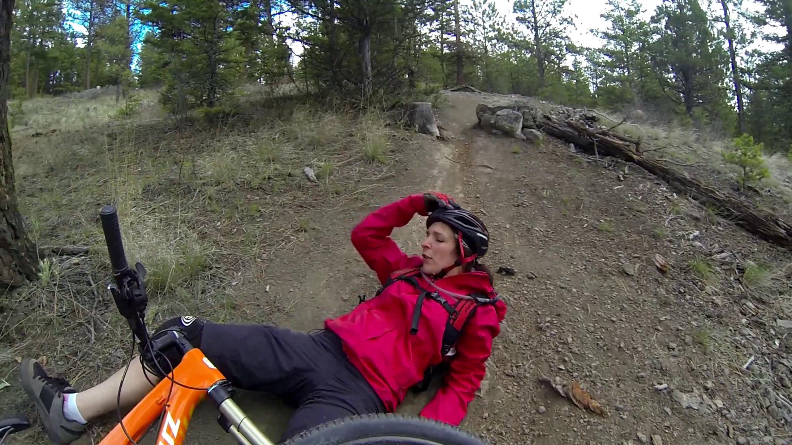 A person wearing a helmet and a red jacket is sitting on the ground beside a mountain bike on a dirt trail in a forested area. The individual appears to be recovering from a fall, with one hand on their forehead and a concerned expression. Surrounding them are trees and rocks, indicating a natural outdoor setting. Smith Creek mountain bike trail.