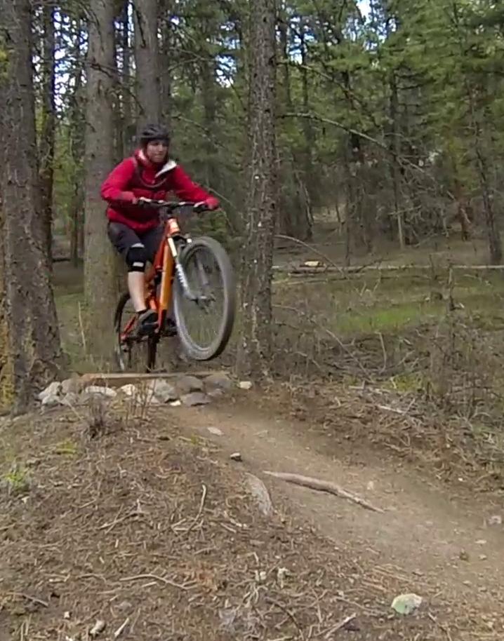 A mountain biker in a red jacket performs a jump on a dirt trail surrounded by trees in a forested area. The bike is airborne, showcasing skill and agility amid a natural setting. Smith Creek mountain bike trail.