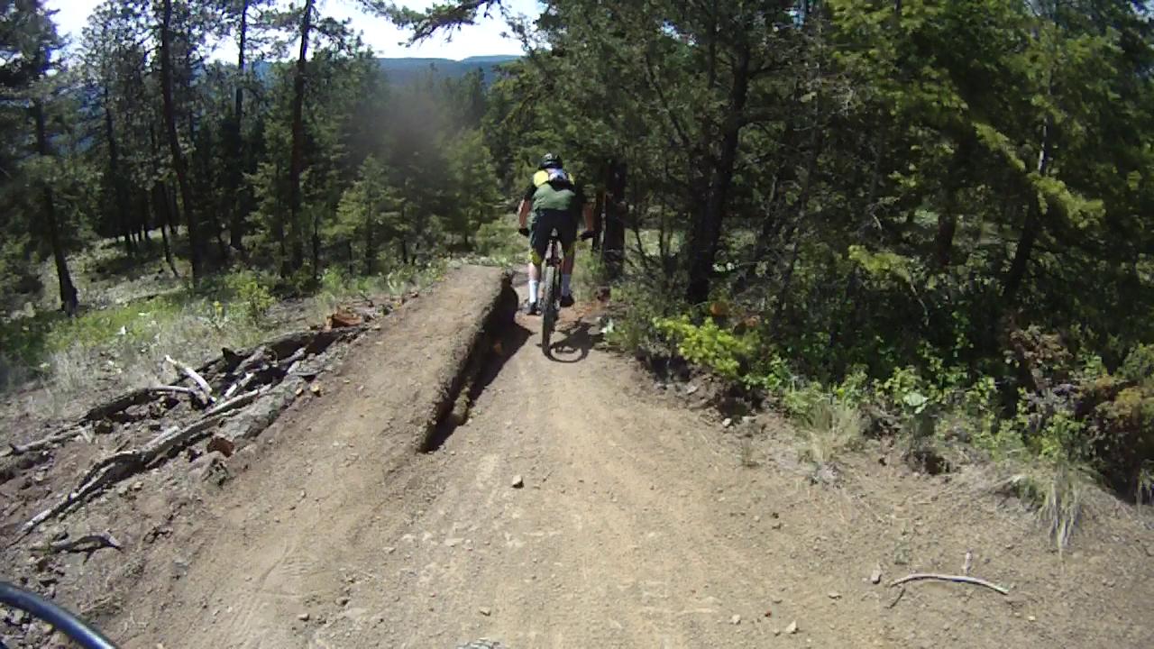 A mountain biker riding on a dirt trail surrounded by trees, approaching a narrow section of the path with a drop-off on one side. The scene captures a sunny day in a forested area, emphasizing the outdoor adventure of mountain biking. Smith Creek mountain bike trail.