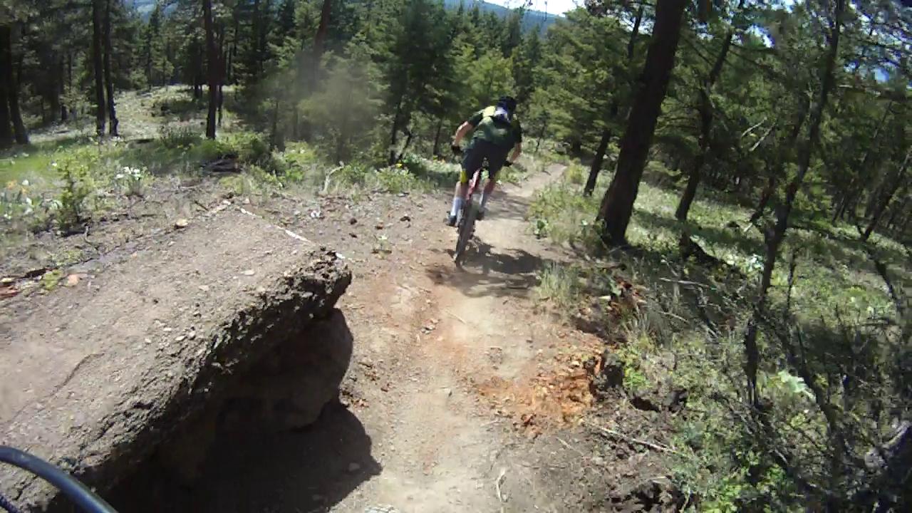 A mountain biker navigating a dirt trail surrounded by trees in a forested area, with a large rock on the path. The scene captures a sunny day, highlighting the greenery and rugged terrain typical of outdoor biking trails. Smith Creek mountain bike trail.