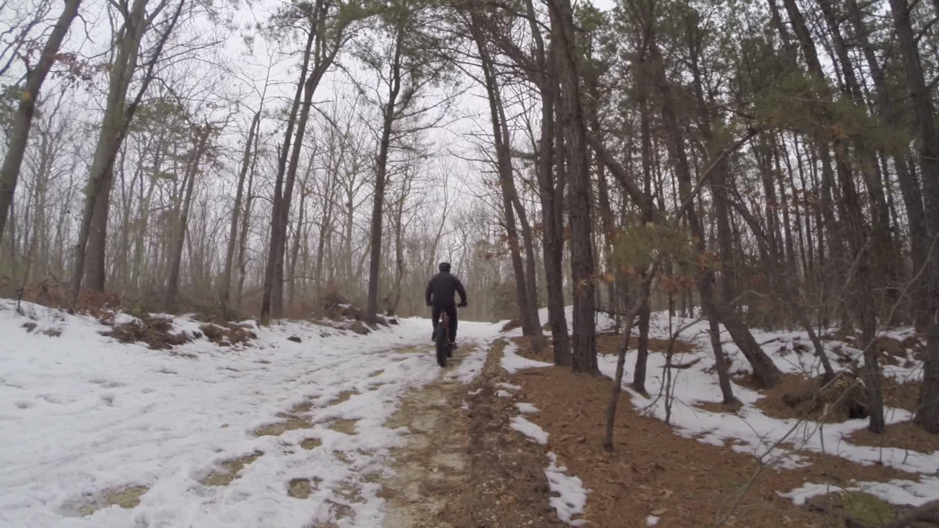 A person riding a mountain bike along a snow-covered trail in a forested area, surrounded by bare trees and patches of snow on the ground. The scene is overcast, indicating a chilly, wintry atmosphere. Allaire State Park mountain bike trail.