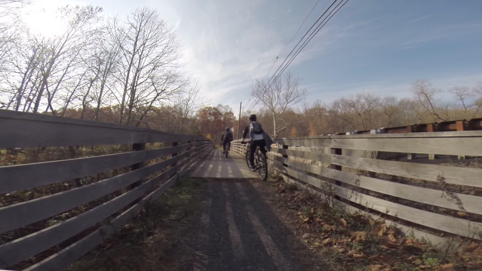 A scenic view of a bike path in a wooded area during autumn, with two cyclists riding along the path. The path is bordered by wooden railings, and the background features bare trees and a clear sky. Sunlight casts shadows on the ground, creating a peaceful outdoor atmosphere. Chimney Rock mountain bike trail.