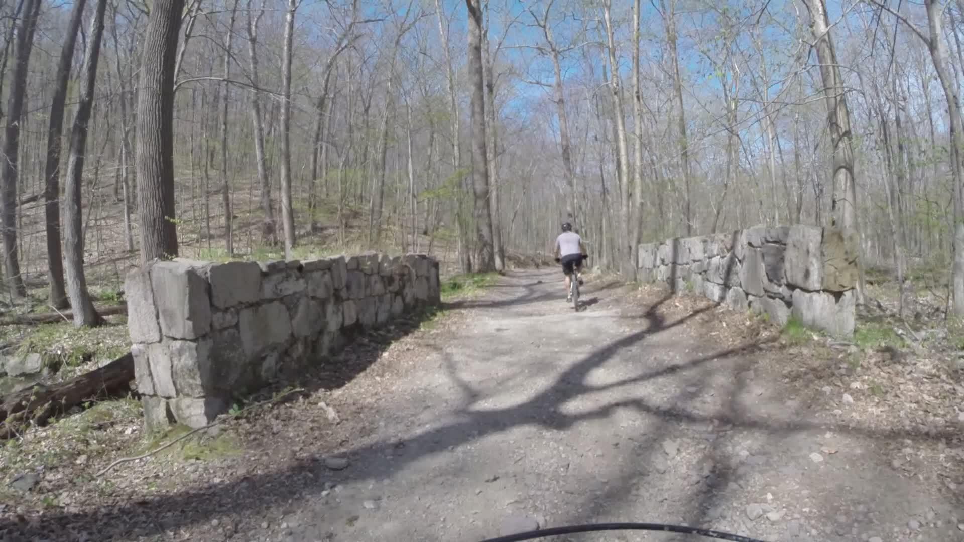 A cyclist riding on a dirt path through a wooded area, with bare trees and stone walls lining the trail. The scene is set under a clear blue sky, indicating a sunny day in early spring. Ringwood Skylands Manor mountain bike trail.