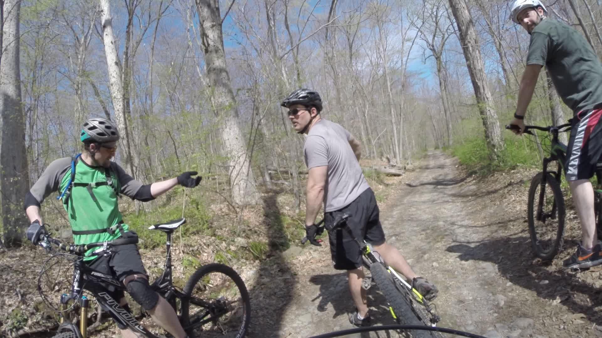 Group of three mountain bikers taking a break on a dirt trail surrounded by trees. One cyclist is pointing while sitting on his bike, another is looking back from his bike, and the third is facing the camera. The scene is set in a wooded area with clear blue skies. Ringwood Skylands Manor mountain bike trail.