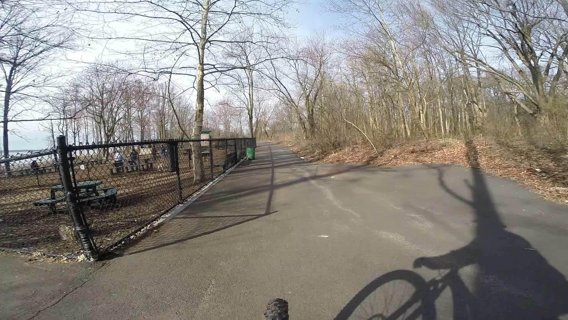 A view of a paved bike path winding through a wooded area, with trees on either side and a fenced area visible to the left. The scene captures shadows from the bike and surrounding trees, with a few picnic tables and a green trash can in the fenced area. Wolfes Pond park mountain bike trail.