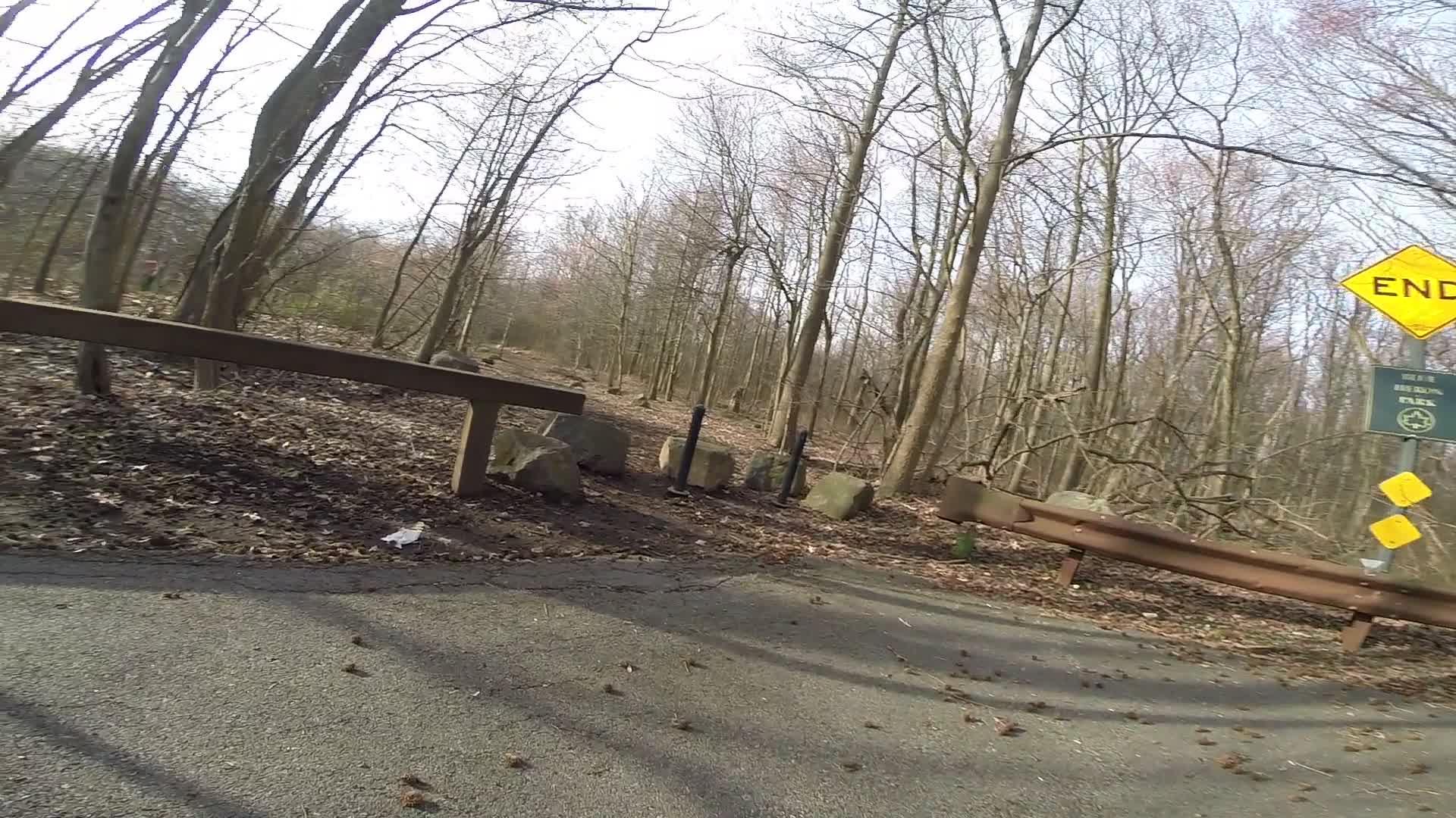 A wooded area with bare trees in a park, featuring a gravel path and a wooden barrier. A yellow "END" sign is positioned near the end of the path, with additional signage visible. Brown leaves are scattered on the ground, indicating early spring or late autumn. Trails seperated by streets mountain bike trail.