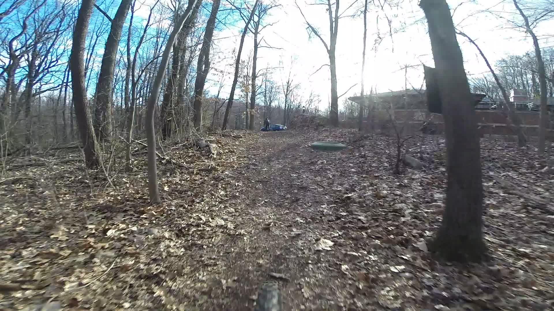 A winding dirt trail through a wooded area, covered in fallen leaves, with trees lining both sides. In the background, there's a person near a blue object on the ground, with a residential structure partially visible through the trees. The sky is bright with scattered clouds. Richmond Avenue and Forest Hill road mountain bike trail.