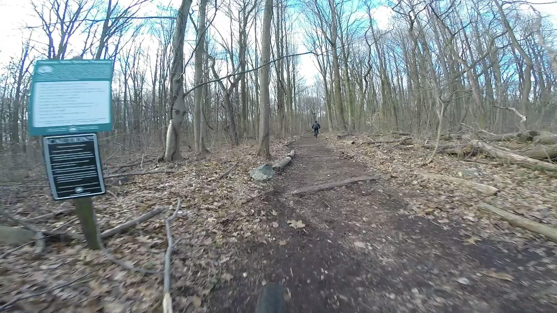 A winding dirt path through a forested area, with bare trees and fallen leaves lining the trail. A person in the distance is walking along the path, and there are signs visible on the left side of the image providing cautionary information about the trail. The scene is set under a cloudy sky. Richmond Avenue and Forest Hill road mountain bike trail.